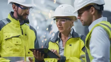 Three workers in safety gear discuss a tablet in an industrial setting.