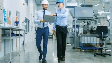 Two people walking in a factory, one holding a laptop.