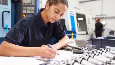 Individual writing at an industrial workstation with CNC machines in the background.