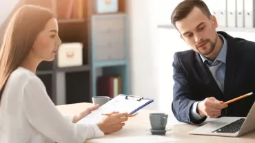Two people at a desk having a discussion, one gesturing towards a laptop.